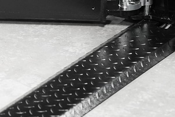 Nahaufnahme eines diagonal verlaufenden schwarzen Riffelblech-Streifens mit diamantförmiger Struktur, am oberen Rand sichtbare Metallmechanik auf hellem glattem Untergrund