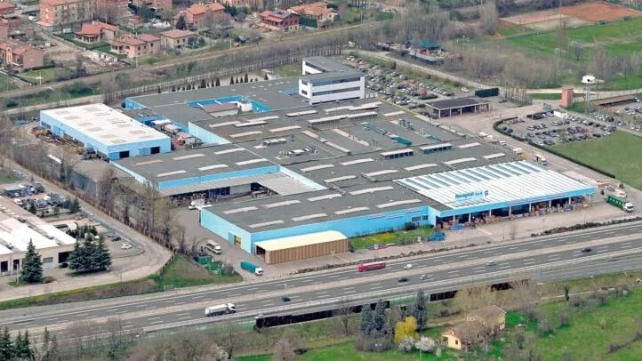 Luftaufnahme eines großen blau-grauen Industriekomplexes mit verbundenen Hallendächern, Parkplätzen, Lkw an einer vierspurigen Schnellstraße und umliegenden Wohnhäusern und Feldern