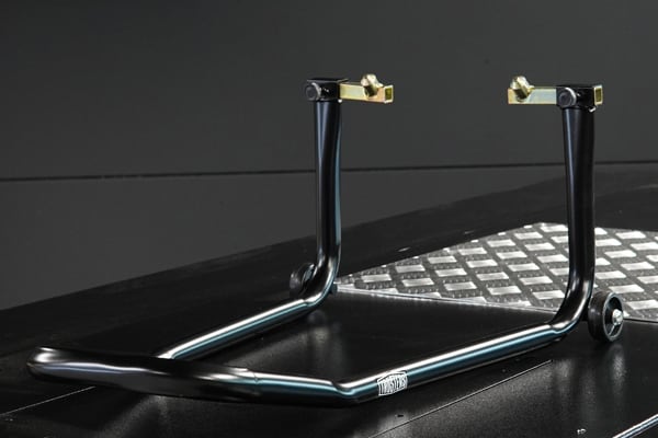 Black steel motorcycle rear paddock stand with U-shaped base, two vertical supports topped by brass-colored adjustable fittings and small wheels, set on a black surface next to a diamond-plate metal panel