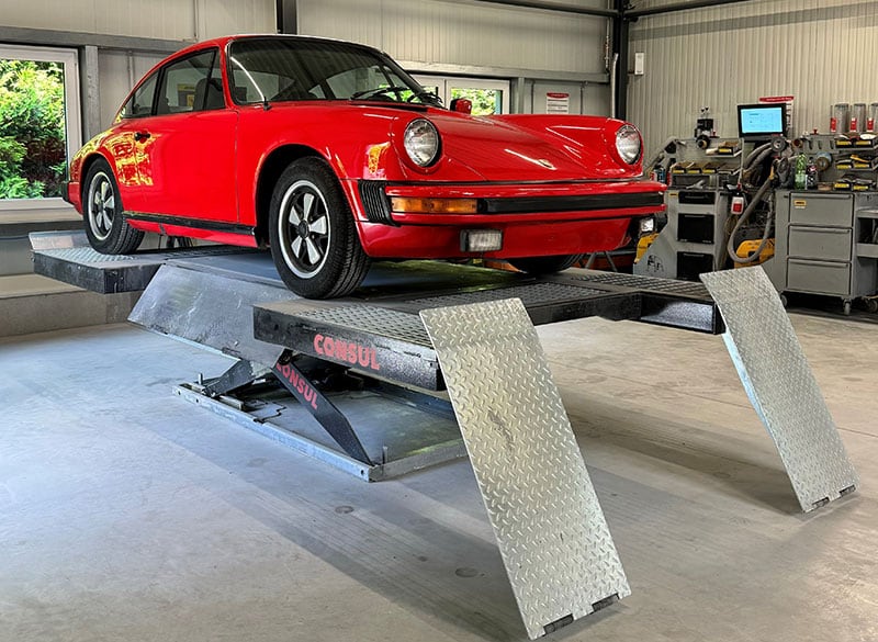 Roter klassischer Porsche 911 auf Hebebühne in Werkstatt, Metallrampen vor dem Fahrzeug, Werkzeugschränke und Diagnosegerät im Hintergrund, Betonboden und Fensterlicht