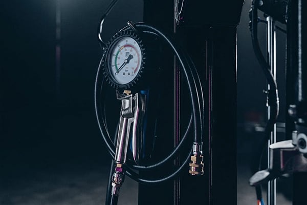 Manometer mit aufgewickeltem Luftschlauch und Metallanschluss, an schwarzer Werkstattsäule hängend, Skala mit farbigen Bereichen, dunkle Garage im Hintergrund