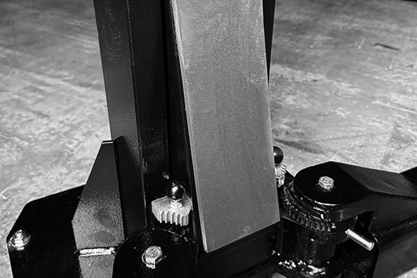 Nahaufnahme einer metallischen Maschinenbasis mit vertikalem rechteckigen Stützpfosten, sichtbaren Zahnrädern, Schrauben und Befestigungsplatten auf Betonboden, monochromatisch