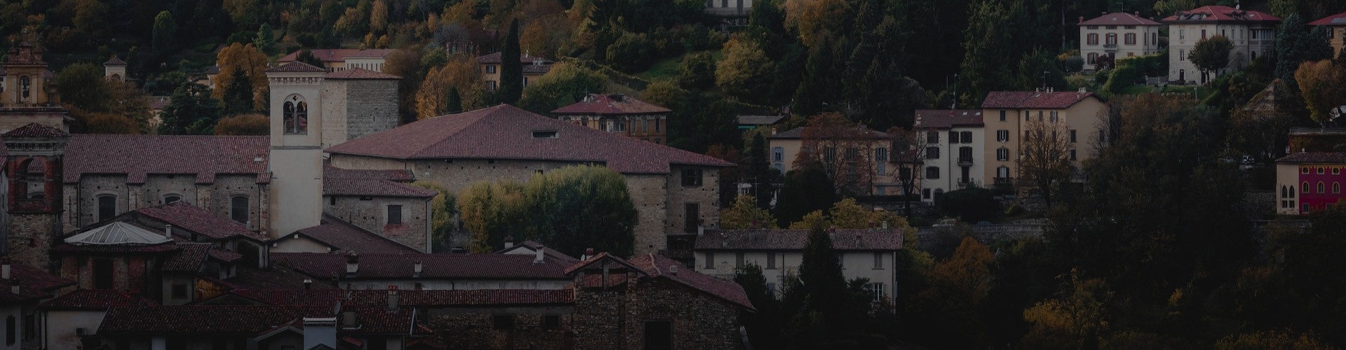 Panoramablick auf eine hügelige Kleinstadt mit alten Steinhäusern und roten Ziegeldächern, einem Kirchturm, herbstlich gefärbten Bäumen und dicht aneinandergereihten Wohnhäusern