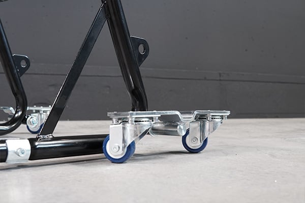 Close-up of two blue rubber caster wheels on silver metal brackets attached to a black painted tubular frame, resting on a smooth gray concrete floor with a gray wall background