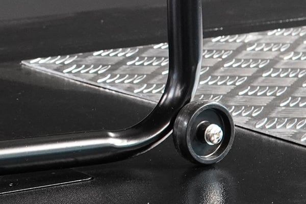 Close-up of a small black caster wheel attached to a curved black metal frame, resting on a glossy black surface beside a raised diamond-pattern metal plate