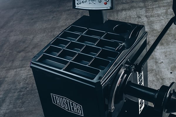 Schwarze Industriemaschine mit mehreren rechteckigen Ablagefächern, Display/Bedienfeld oben, seitlichem Hebel und Handrad, Aufschrift TRUSTERS auf der Front, steht auf grauem Betonboden