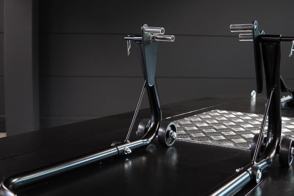 Caballete de moto metálico negro en primer plano sobre suelo de taller oscuro, con una placa de metal cuadriculada antideslizante al fondo