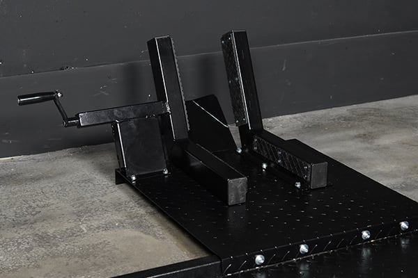 Calzo metálico negro para rueda de motocicleta sobre placa perforada, con dos soportes verticales, un bloque longitudinal y una manija, apoyado en suelo de hormigón frente a pared gris