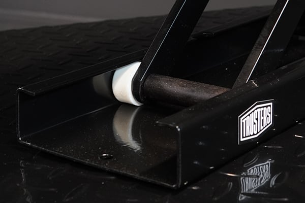 Primer plano de soporte metálico negro con rodillo blanco y barra acolchada, logo TRUSTERS en el lateral, sobre superficie negra brillante con reflejo