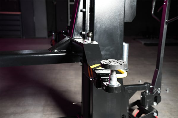 Close-up of adjustable threaded support pad with rubber top on a black metal lift arm in a dimly lit garage or workshop, concrete floor and caster wheel visible