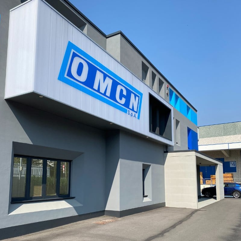 Industrie-/Bürogebäude mit grauer Fassade und großem blau-weißem OMCN S.P.A.-Logo, überdachter Eingang, Fensterfront, Parkplatz mit geparktem Auto, klarer blauer Himmel