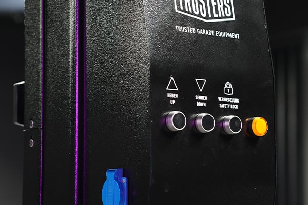 Close-up of black garage equipment control panel with three silver push buttons labeled UP, DOWN and SAFETY LOCK, an illuminated yellow button, a blue power socket, purple edge trim and textured metal surface
