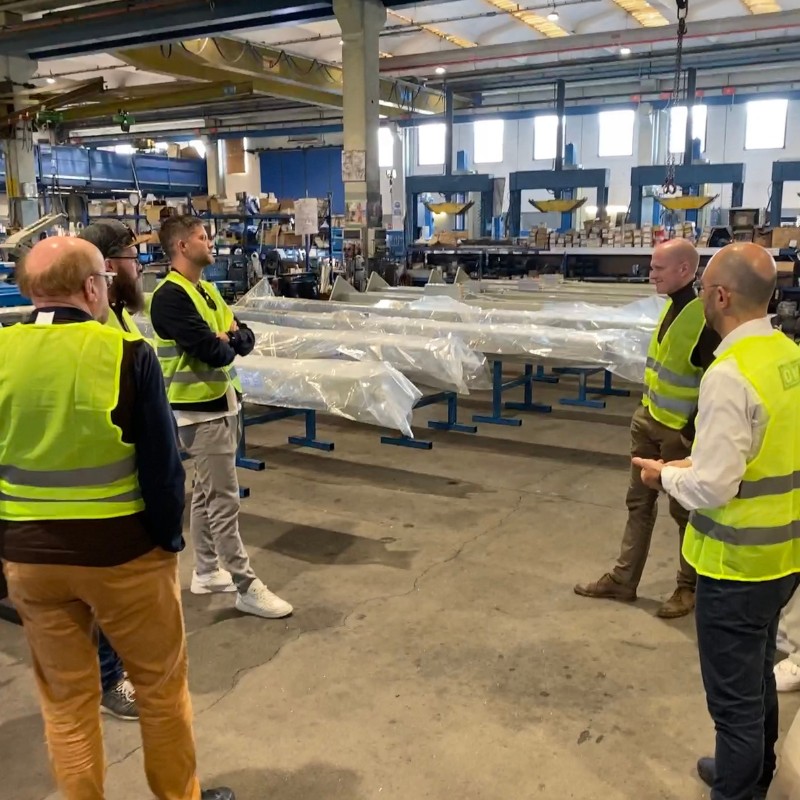 Mehrere Personen in gelben Warnwesten diskutieren in einer Werkhalle neben auf Gestellen verpackten langen Metallträgern, im Hintergrund sind Maschinen, Regale und Deckenbeleuchtung zu sehen