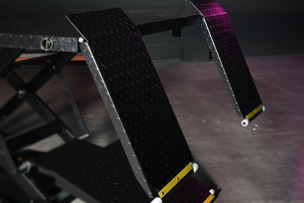 Close-up of two black diamond-plate metal ramps with yellow end caps attached to a scissor lift platform, angled downward toward a concrete floor with purple accent lighting