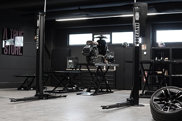 Dunkle, moderne Autowerkstatt mit zwei Hebebühnenpfeilern, mittig erhöhter Motorradmotor auf Arbeitspodest, Werkzeugschränken an der Wand, Reifen im Vordergrund und leuchtender Wandbeschriftung