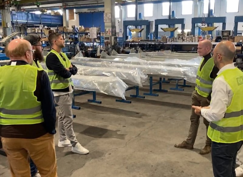 Fünf Personen in gelben Warnwesten stehen in einer Werkshalle im Kreis und besprechen verpackte lange Metallprofile auf blauen Gestellen, im Hintergrund Regale und Maschinen