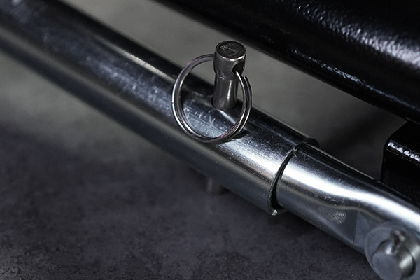 Nahaufnahme eines verchromten Metallrohrs mit eingestecktem Sicherungsstift und rundem Splint-Sicherungsring, Teil einer klappbaren Metallkonstruktion, oberhalb schwarzer Polsterung und darunter grauer Boden