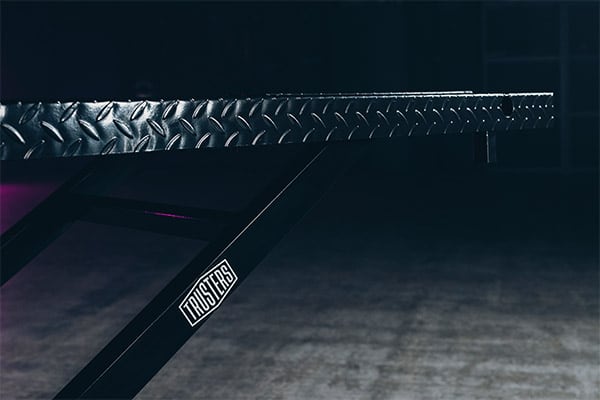 Close-up of a diamond-plate metal bench edge and black angled support leg with TRUSTERS logo against a dimly lit gym floor