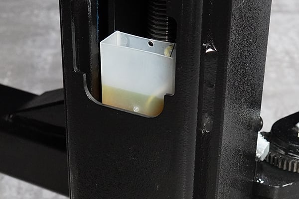 Close-up of a white plastic lubricant reservoir partly filled with yellow fluid mounted inside a black metal vertical support of a mechanical lift, with a visible threaded rod and gear mechanism at the side