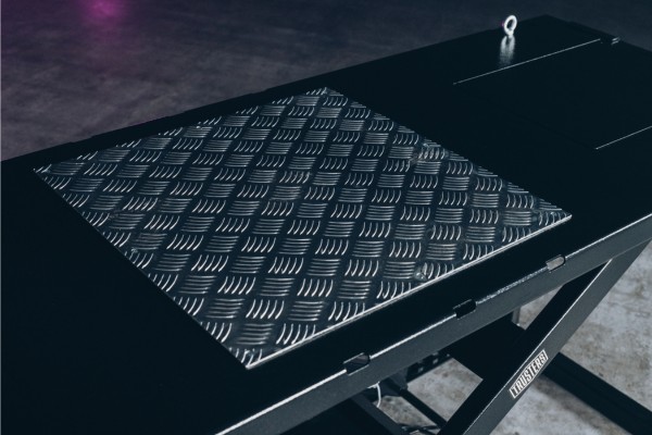 Placa metálica antideslizante con patrón de rombos sobre una plataforma negra de mesa elevadora tipo tijera, detalle de textura brillante, puntos de anclaje visibles y suelo de hormigón con leve reflejo púrpura