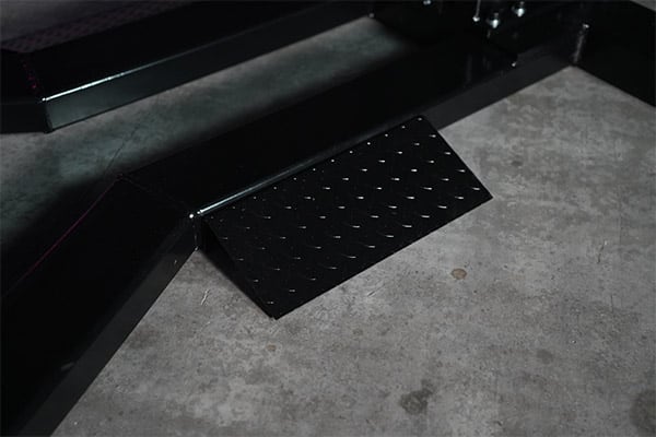 Black diamond-pattern steel footplate mounted on a black metal frame, positioned on a worn concrete floor with small stains