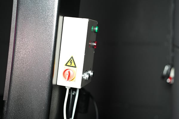 Caja eléctrica blanca montada en un bastidor oscuro con interruptor rojo giratorio, dos indicadores luminosos (verde y rojo), símbolo de alto voltaje en amarillo y varios cables salientes