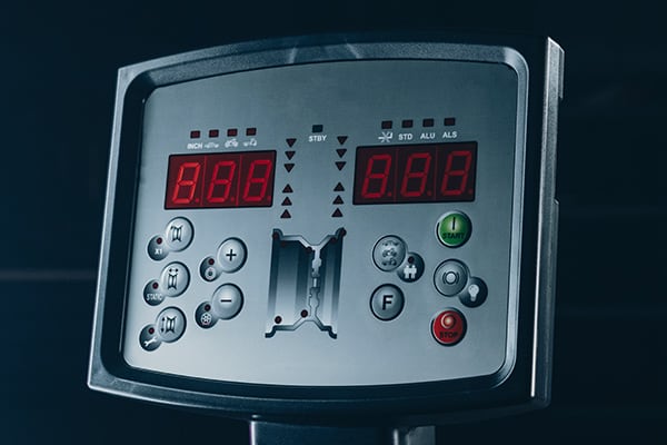Maschinenbedienfeld mit zwei roten 7-Segment-Anzeigen, zentraler Einstecköffnung und Pfeilanzeigen, mehrere runde Funktionstasten mit Symbolen, grüne Starttaste und rote Stopptaste, metallisches Gehäuse auf Ständer vor dunklem Hintergrund