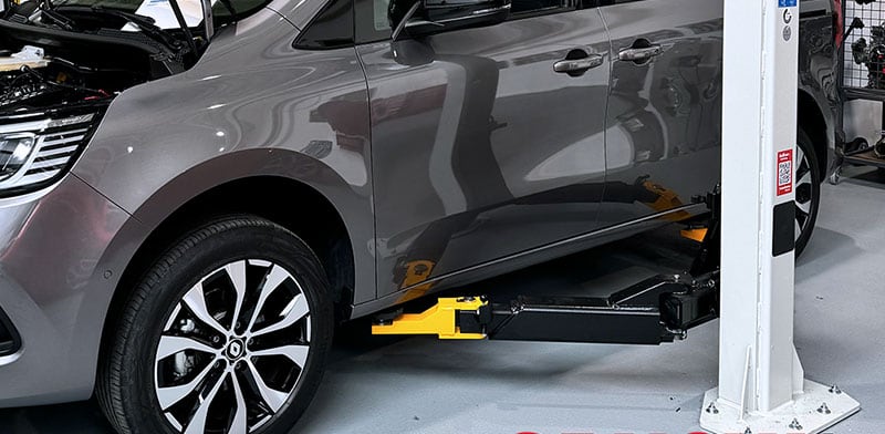 Graues Auto auf Werkstatt-Hebebühne, Nahaufnahme der vorderen linken Seite mit Rad/Alufelge, gelb-schwarzer Hebearm unter dem Fahrzeug, weiße Säule der Hebebühne und glatter Werkstattboden im Hintergrund