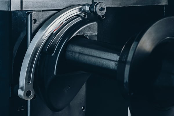Nahaufnahme eines gebogenen Metallhebels an einer Maschine mit zylindrischer Achse und runder Rolle, sichtbaren Schrauben und dunklen mattierten Metalloberflächen