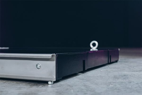 Nahaufnahme einer niedrigen Metallplattform oder Bühnenrampe mit schwarzer Seitenverkleidung, silbernem Rahmen, sichtbarer Schraube und Öse auf grauem Betonboden vor dunklem Hintergrund