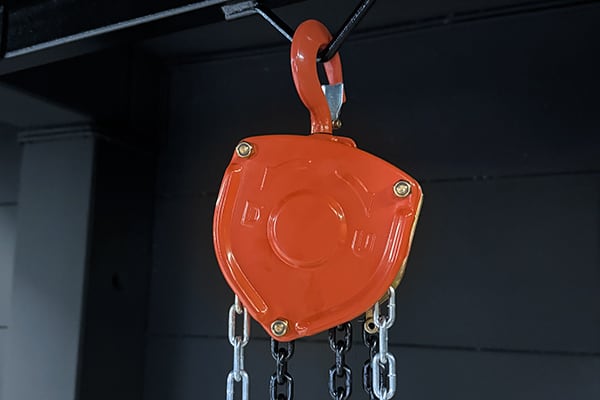 Polipasto manual naranja colgando de un gancho con cadenas metálicas, detalle del mecanismo contra un fondo oscuro industriel