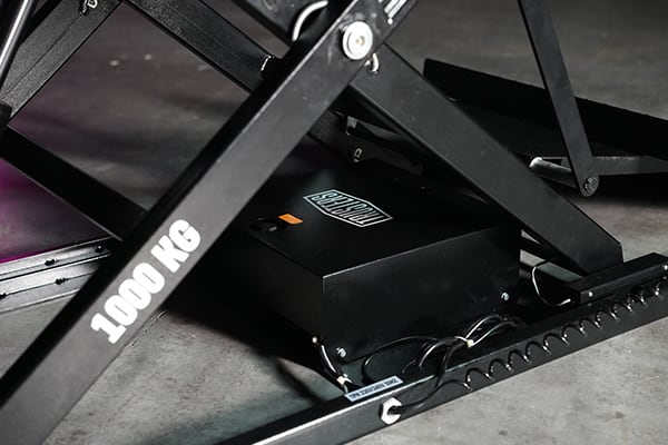 Close-up of a black scissor lift mechanism on a concrete floor showing a 1000 KG label on the frame, a black control box with cables and a small orange indicator light