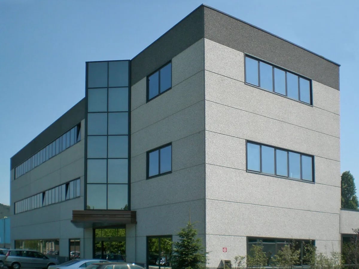 Dreigeschossiges modernes Bürogebäude mit hellgrauer Fassade, vertikalem Glas-Eingangsbereich, rechteckigen Fenstern, geparkten Autos und kleinen Bäumen vor klarem blauem Himmel