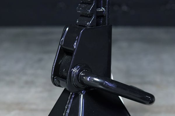 Nahaufnahme eines schwarz lackierten Metall-Unterstellbocks mit Ratschenstange und eingestecktem Sicherungsbolzen, auf leicht verschwommenem Betonboden