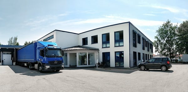 Zweistöckiges weißes Büro- und Lagergebäude mit blauen Fensterrahmen, überdachtem Eingang, blauem Lkw links, schwarzem Pkw rechts, Fahrrad am Eingangsbereich und großem asphaltiertem Parkplatz unter hellem Himmel