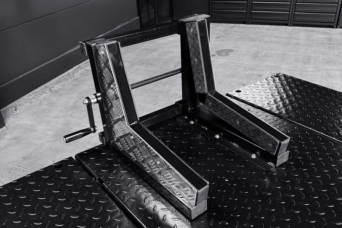 Black painted steel wheel chock and support frame with hand crank mounted on a diamond-plate lift platform, photographed in a garage with concrete floor and tool cabinets in the background