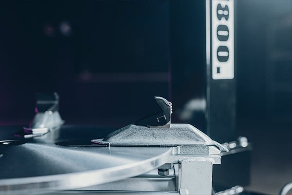 Detalle de la cápsula y aguja de un tocadiscos sobre un disco giratorio metálico, con un soporte vertical numerado desenfocado al fondo