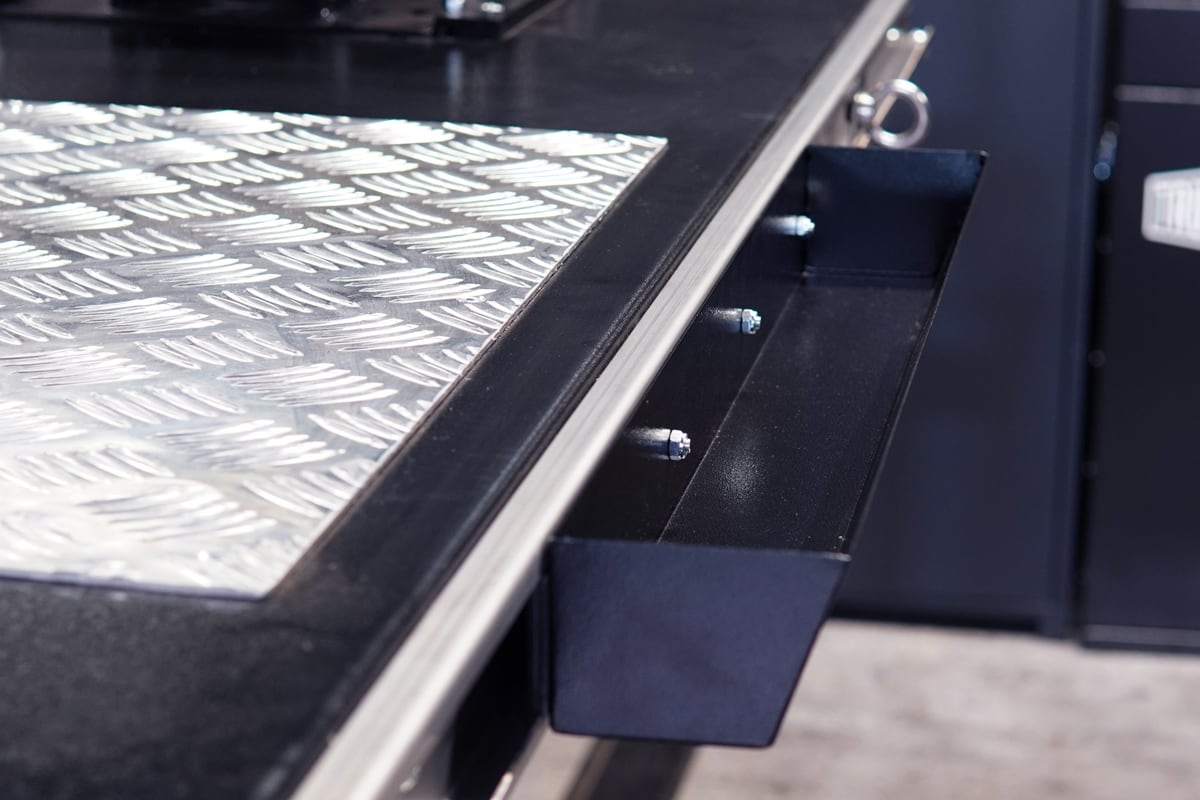 Nahaufnahme einer Metallarbeitsplatte mit rutschfester Riffelblech-Oberfläche, schwarzer Kante und offenem schmalem Werkzeugfach mit sichtbaren Schrauben