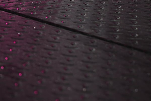 Close-up of dark textured metal surface with repeating small raised heart-shaped studs, a shallow diagonal seam, and soft pink highlights and blurred light reflections