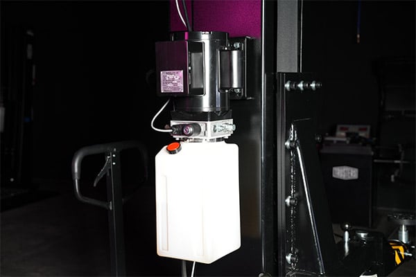White plastic container with orange cap mounted beneath a small electric motor on a black metal industrial machine, showing mounting brackets, bolts and cables against a dim workshop background