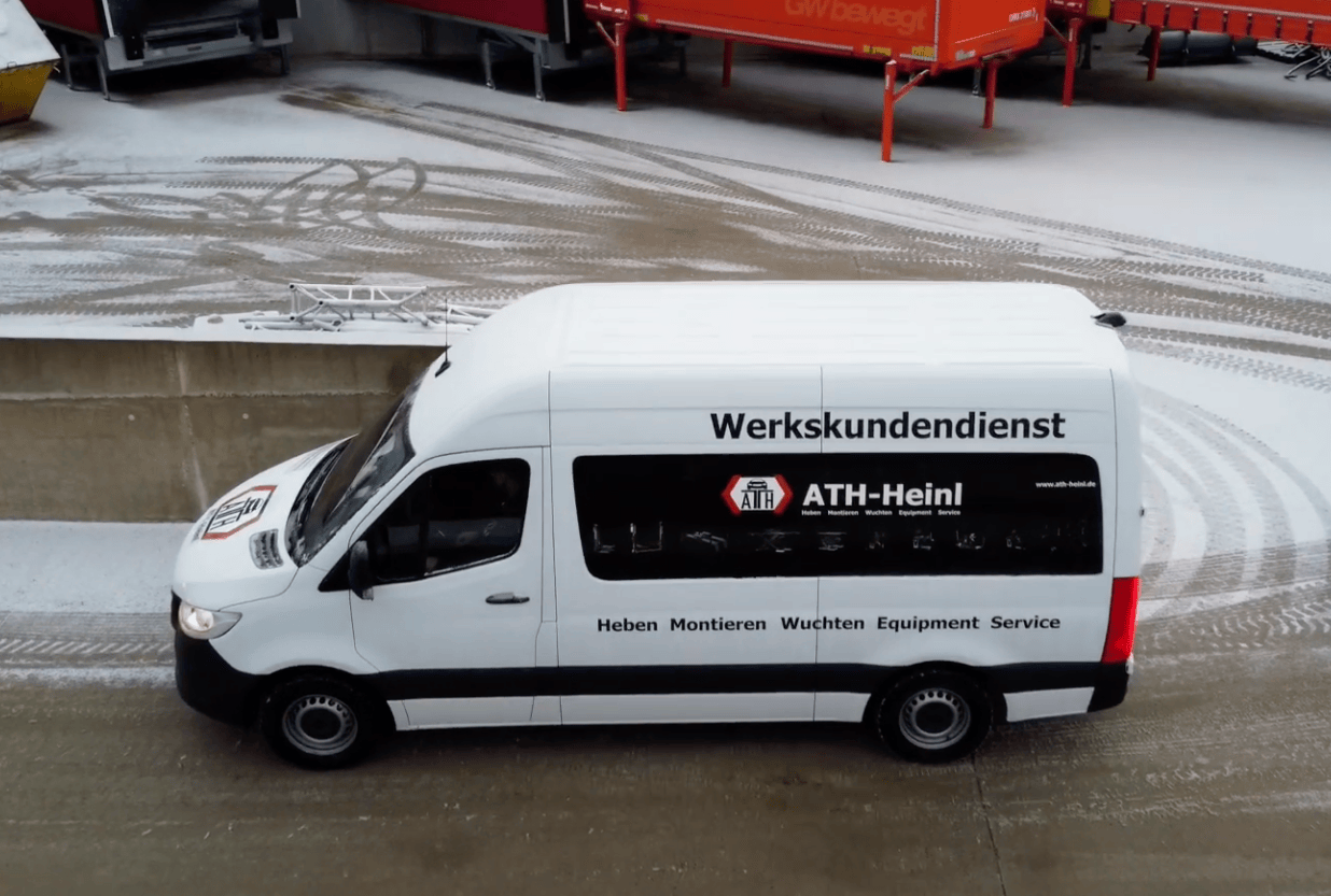 Weißer Transporter mit Aufschrift Werkskundendienst und Firmenlogo ATH‑Heinl, geparkt auf leicht verschneitem Hof mit Reifenspuren; rote Anhänger und Betonmauer im Hintergrund.