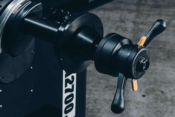 Nahaufnahme einer schwarzen mechanischen Spindel mit zwei ergonomischen Handgriffen und gelben Verriegelungshebeln, montiert an einer Metallmaschine mit weißer Nummernmarkierung und Betonboden im Hintergrund