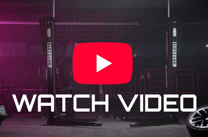 Garage interior with dual car lifts and tire rim, overlaid by large red YouTube play button icon and bold white text WATCH VIDEO, dim purple lighting and faint outlined VIDEO text in background.