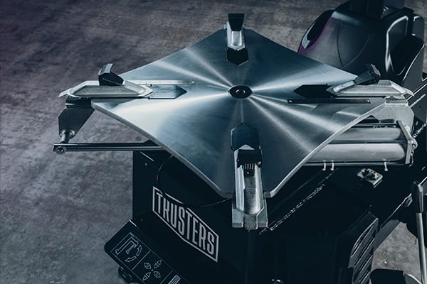 Industriergerät mit quadratischer, gebürsteter Metallplatte und vier verstellbaren Spannarmen auf schwarzer Basis, Vorderseite mit Schriftzug THRUSTERS, grauer Werkstattboden im Hintergrund