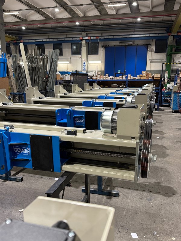 Reihe blauer-beiger Industrieantriebe mit Motoren und Riemenscheiben in einer Werkhalle, Regale und Kartons im Hintergrund, hohe Decke mit Beleuchtung