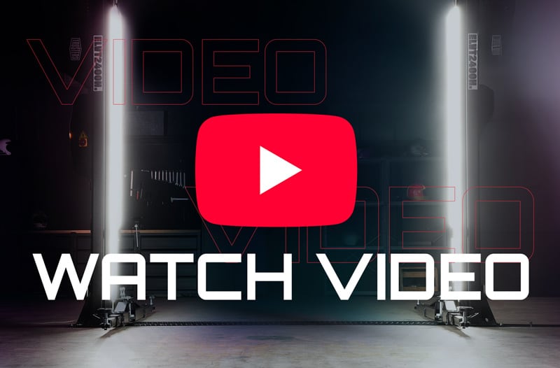 Dark studio with a large red YouTube play button centered above bold white text WATCH VIDEO, flanked by two vertical bright light tubes and faint equipment visible in the background