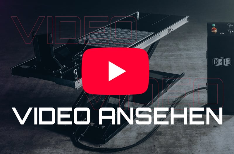 Schwarze Scherenhebebühne mit geriffelter Metallplattform auf Betonboden, Hydraulikschläuche und Steuerbox rechts mit TRUSTERS-Logo, überlagert von großem roten YouTube-Play-Symbol und weißem Schriftzug VIDEO ANSEHEN