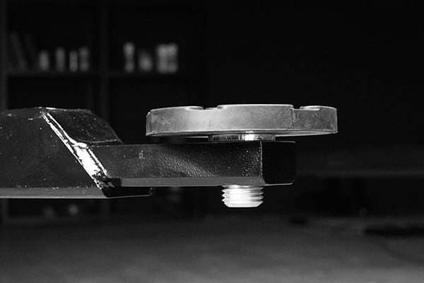 Close-up of a black painted metal arm supporting a circular silver metal plate with a threaded bolt protruding downward, shallow depth of field and blurred workshop shelving in the background, black-and-white photograph