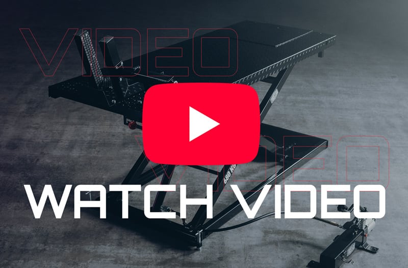 Black hydraulic scissor-lift service table on a concrete floor with a large red YouTube-style play button overlay and bold white text "WATCH VIDEO" across the foreground