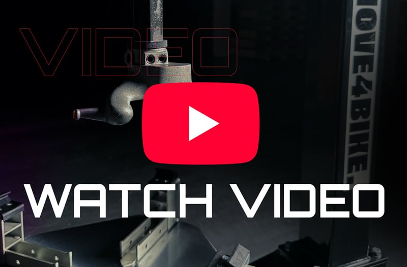 Industrial metal robotic arm and fixtures in a dark workshop background with a large red YouTube play button overlay at center and bold white text WATCH VIDEO across the bottom